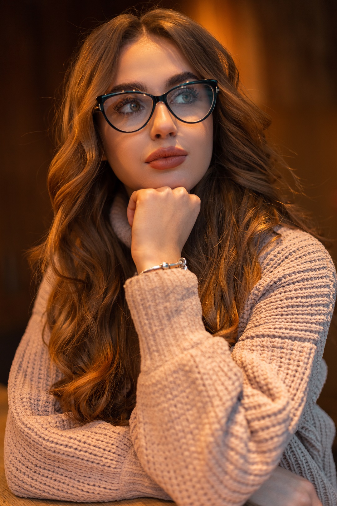 Indoors portrait of a beautiful stylish woman with trendy glasses and curly hairstyle in a fashion beige knitted vintage sweater sits in cafe